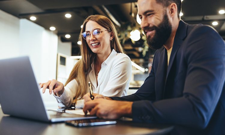 Cheerful businesspeople using a laptop in an office. Happy young entrepreneurs smiling while working together in a modern workspace. Two young businesspeople sitting together at a table.